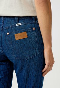 Jeans in denim blu scuro con una texture a motivo. Presentano un'etichetta in pelle con "Wrangler" e dettagli in rame sulle tasche posteriori.