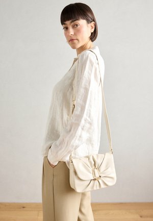 Kobieta o krótkich brązowych włosach, ubrana w luźną białą bluzkę i beżowe spodnie, niosąca na ramieniu kremową torebkę z złotym zapięciem.