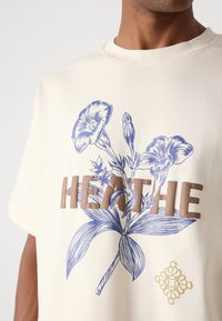 Cremefarvet t-shirt med et blåt blomsterdesign og brun "HEATHE" tekst. Indeholder et lille gult dekorativt element.