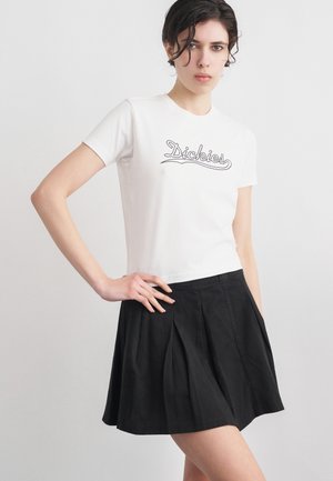 Noor naine lühikeste tumedate juustega, kellel on seljas valge Dickiesi t-särk ja must volditud seelik, poseerib käega puusal ühevärvilise tausta ees.