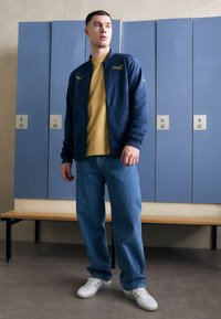 Veste de sport zippée navy avec accents texturés, T-shirt moutarde, jean bleu loose et sneakers blanches, devant des casiers.