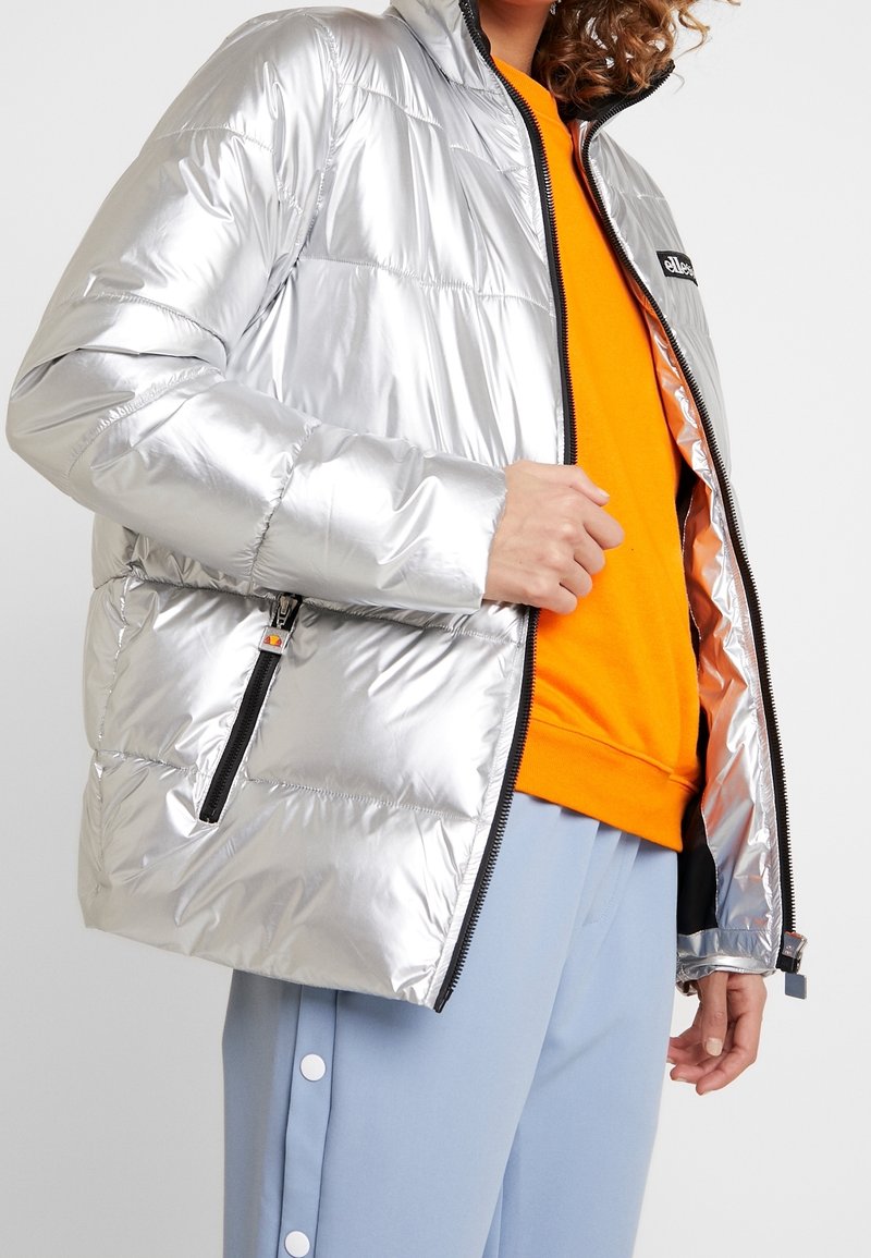 Personne portant une doudoune argentée brillante, un sweat-shirt orange et un pantalon bleu clair avec des boutons blancs, tenant la fermeture éclair de la veste partiellement ouverte.
