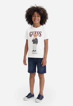 Smilende barn med krøllet hår iført en hvid t-shirt med et bjørnemotiv, mørke denimshorts og marineblå sneakers.