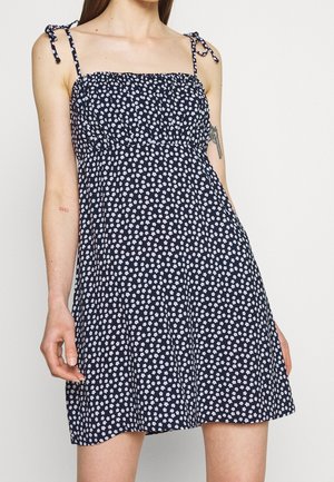 Žena v tmavě modrých bezrukávových šatech s malým bílým květinovým potiskem a zavazovacími ramínky, ukazující tetování na obou pažích.