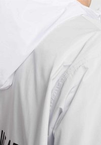 Biała kurtka przeciwdeszczowa z kapturem, gładką fakturą i minimalistycznym designem z akcentowanym logo na plecach.