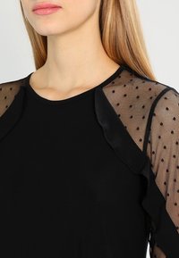 Svart topp med genomskinliga prickiga meshärmar. Halsringningen är rundad, och det finns en volangdetalj på axlarna. Slät tygtextur.