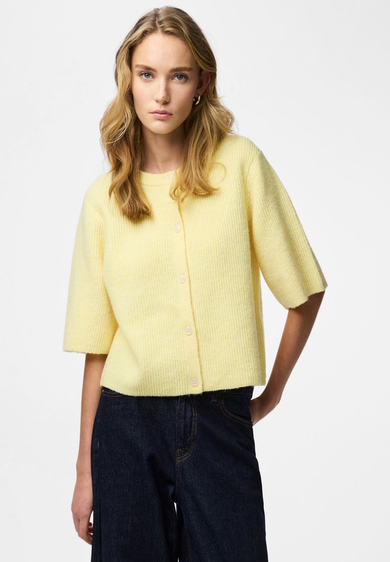 Short-sleeve, cropped cardigan in soft yellow ribbed knit, featuring a round neckline and button closure at the front. Worn with dark jeans.