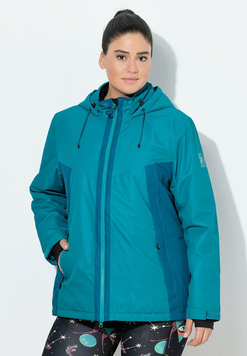 Kvinna klädd i en turkos zip-up jacka med huva och justerbara snören samt mönstrade leggings med himmelska motiv.