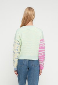 Γυναίκα με μακριά ξανθά μαλλιά, φορώντας πουλόβερ παστέλ με ανάμεικτα ζωικά σχέδια και μπλε τζιν, κοιτώντας προς τα πίσω με φόντο ένα λευκό τοίχο.