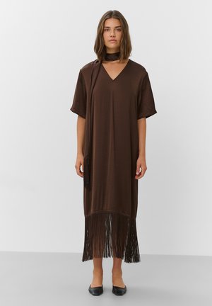 Femme portant une robe midi marron à col en V avec manches courtes, ourlet à franges, foulard assorti et chaussures plates noires, debout sur fond blanc.