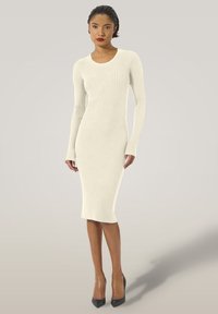 Abito midi in maglia color crema con texture a coste, maniche lunghe, scollo rotondo e silhouette aderente, abbinato a décolleté nere con tacco a spillo.