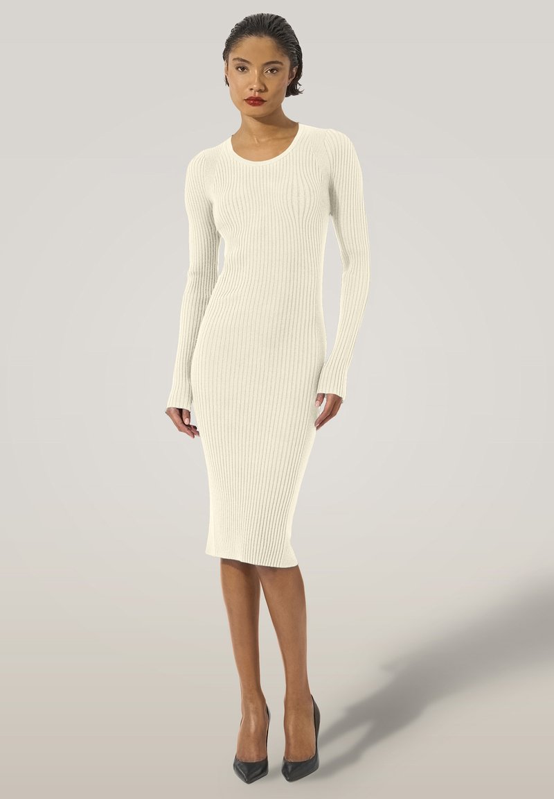 Abito midi in maglia color crema con texture a coste, maniche lunghe, scollo rotondo e silhouette aderente, abbinato a décolleté nere con tacco a spillo.