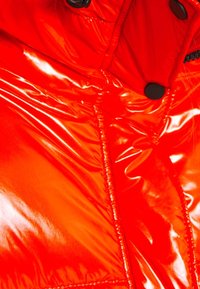 Doudoune rouge vif et brillante avec une texture brillante, présentant des boutons en métal noir et des détails de couture le long des coutures.