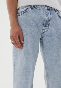 Světle modré džínové kalhoty s volnějším střihem, kontrastním prošíváním, zapínáním na knoflík a předními kapsami. Látka má vybledlou texturu.