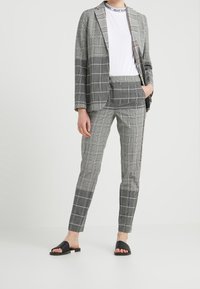 Blazer a quadri grigi e pantaloni slim-fit coordinati, abbinati a una t-shirt bianca e sandali neri aperti. Il tessuto appare liscio e leggero.