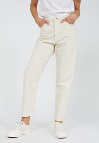 Jeans de color beige claro, de tiro alto, con corte recto, diseño de cinco bolsillos y cierre de botones. Combinados con zapatillas blancas.