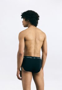 Čierne bavlnené nohavičky s elastickým pásom, na ktorom je logo "Calvin Klein", zobrazené na modeli zo zadnej perspektívy.
