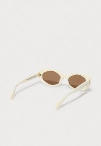 Óculos de sol ovais off-white com lentes castanhas, pousados sobre uma superfície branca, angulados para mostrar o interior das hastes e das dobradiças.