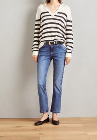 Randig krämfärgad och svart stickad tröja med V-ringning, kombinerad med ljusblå raka jeans och svarta lägenheter, mot en neutral bakgrund.