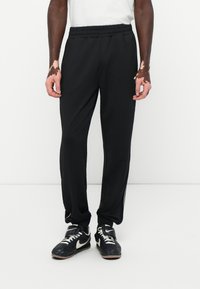 Sorte træningsbukser med elastisk talje og sidelister, båret af en person der står, sammen med sorte og hvide sneakers.