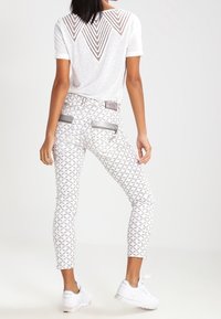 Camisa blanca con un respaldo de patrón triangular, combinada con jeans gris claro que presentan un estampado geométrico, bolsillos con cremallera y corte hasta el tobillo.