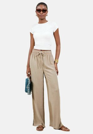 Frau trägt beige breite Hosen, weißes kurzes T-Shirt, braune Sandalen, Sonnenbrille mit Schildpattfassung, goldenes Manschettenarmband und hält eine blaue Fransenhandtasche.