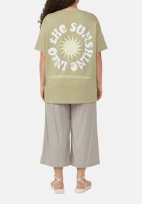 Hellgrünes, übergroßes T-Shirt mit weißem Text und einer Sonnenillustration auf der Rückseite, kombiniert mit grauen, weiten Hosen und hellen Sandalen.