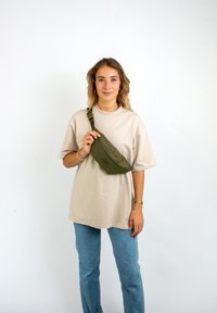 Olivgrüne Bauchtasche aus strapazierfähigem Stoff, gebogene Form, mit einem Reißverschlussfach, getragen über einem lockeren beigen T-Shirt und hellblauen Jeans.
