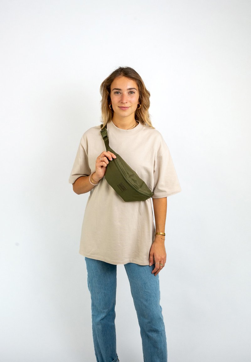 Olivgrüne Bauchtasche aus strapazierfähigem Stoff, gebogene Form, mit einem Reißverschlussfach, getragen über einem lockeren beigen T-Shirt und hellblauen Jeans.