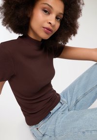 Brązowa, prążkowana bluzka na krótki rękaw z wysokim kołnierzem, w zestawieniu z jasnoniebieskimi dżinsami o wysokim stanie. Gładka tekstura, dopasowany krój.