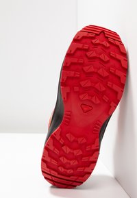 Black athletic shoe with a red rubber sole, featuring deep treads and textured patterns for traction. Side branding visible.
