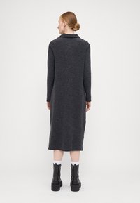 Rochie tricotată lungă, gri închis, cu guler, mâneci până la podea și croi drept. Asortată cu cizme cu platformă negre. Fără modele vizibile.