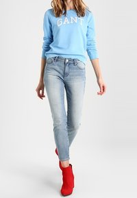 Jasnoniebieska bluza z napisem "GANT" w białym kolorze, w połączeniu z jasnoniebieskimi dżinsami. Widać czerwone botki do kostki oraz długie włosy.