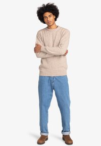Beige gebreide trui met een ronde hals, een gestructureerd oppervlak en geribde manchetten, gecombineerd met lichtblauwe relaxed-fit jeans en bruine laarzen.
