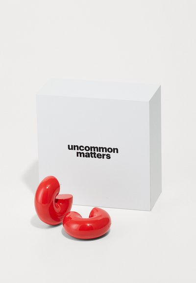 Rote, glänzende Keramikohrringe mit einem geschwungenen Design, kombiniert mit einer weißen Box, die den Text "uncommon matters" in fettem Schwarz zeigt.