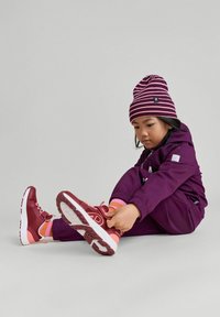 Dziecko w purpurowym kombinezonie narciarskim i w pasiastej purpurowej czapce, siedzi i poprawia czerwone i różowe buty sportowe z białymi podeszwami.