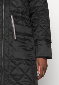 Chaquetón acolchado negro con cierre de cremallera, bolsillo lateral con tira de tirador y detalles de puños acanalados. Acabado suave y resistente al agua.