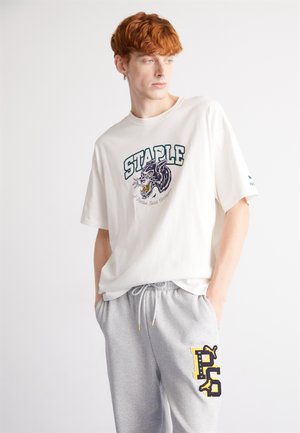 Weißes T-Shirt mit kurzen Ärmeln, das einen Grafikdruck mit einem Drachen und dem Wort "STAPLE" zeigt. Kombiniert mit grauen Jogginghosen, die ein gelbes Logo aufweisen.