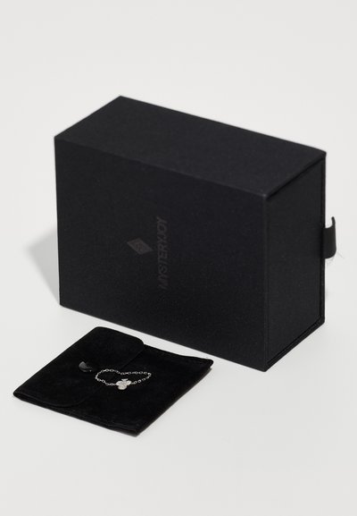 Caja de joyas negra con un acabado mate, con un logo en el lado, junto a una bolsa de terciopelo negro que contiene una pulsera de cadena de plata.