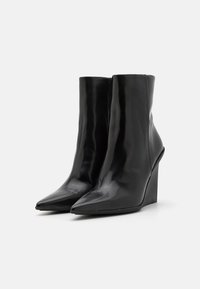 Une paire de bottes noires brillantes à bout pointu et talons compensés sur un fond blanc uni.