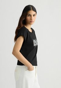 Top noir à manches courtes avec un décolleté carré, ornée d'un design argenté brillant sur le devant. Associé à un pantalon blanc texturé.