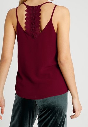 Woman wearing a sleeveless burgundy top with lace and button details on the back, paired with dark velvet pants.