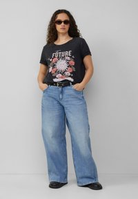 Zwart grafisch T-shirt met florale print met de tekst "De Toekomst," gecombineerd met lichtblauwe wijdleg jeans, een zwarte riem en zwarte schoenen.
