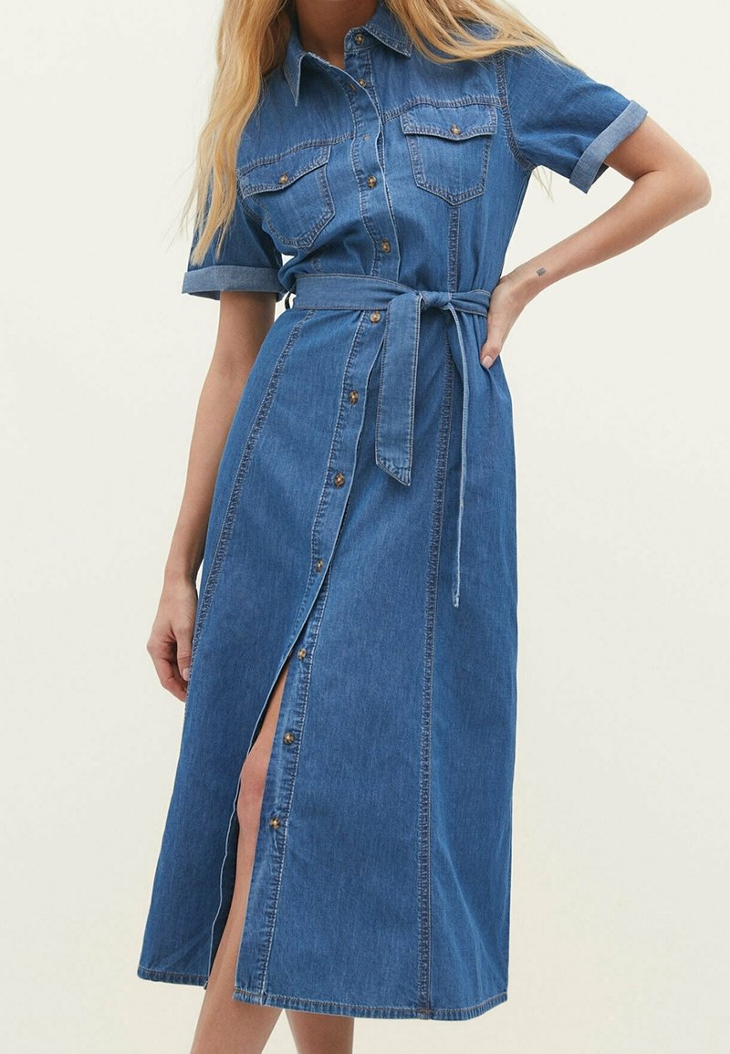 Femme portant une robe midi en denim bleu avec manches courtes, boutonnage devant, poches poitrine et ceinture nouée à la taille, debout avec la main sur la hanche.