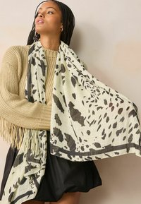 Écharpe légère à motifs en crème avec des taches abstraites noires. Présente une texture douce, des bords frangés et un design fluide.