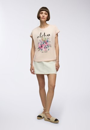 T-shirt rosa chiaro a maniche corte con testo "aloha" e grafica floreale; abbinato a una mini gonna di denim bianca; modella che indossa espadrillas.