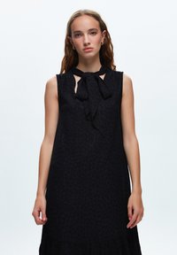 Svart ärmlös klänning med en rosett vid halsen, subtilt texturerat mönster och en utställd nederdel. Har utskärningar vid axlarna.