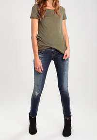 Camiseta de manga corta en verde oliva con un corte relajado, combinada con jeans ajustados de color azul oscuro que presentan detalles desgastados y botas negras de tobillo.