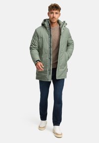 Cappotto verde imbottito con cappuccio, chiusure con zip e bottoni a pressione, indossato sopra un maglione marrone chiaro, abbinato a jeans scuri e sneakers chiare.