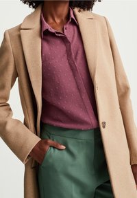 Beige villakangastakki, jossa on tikkulaput, yhdistetty burgundinväriseen teksturoituun paitaan ja vihreisiin housuihin. Takissa on napitus ja taskut.